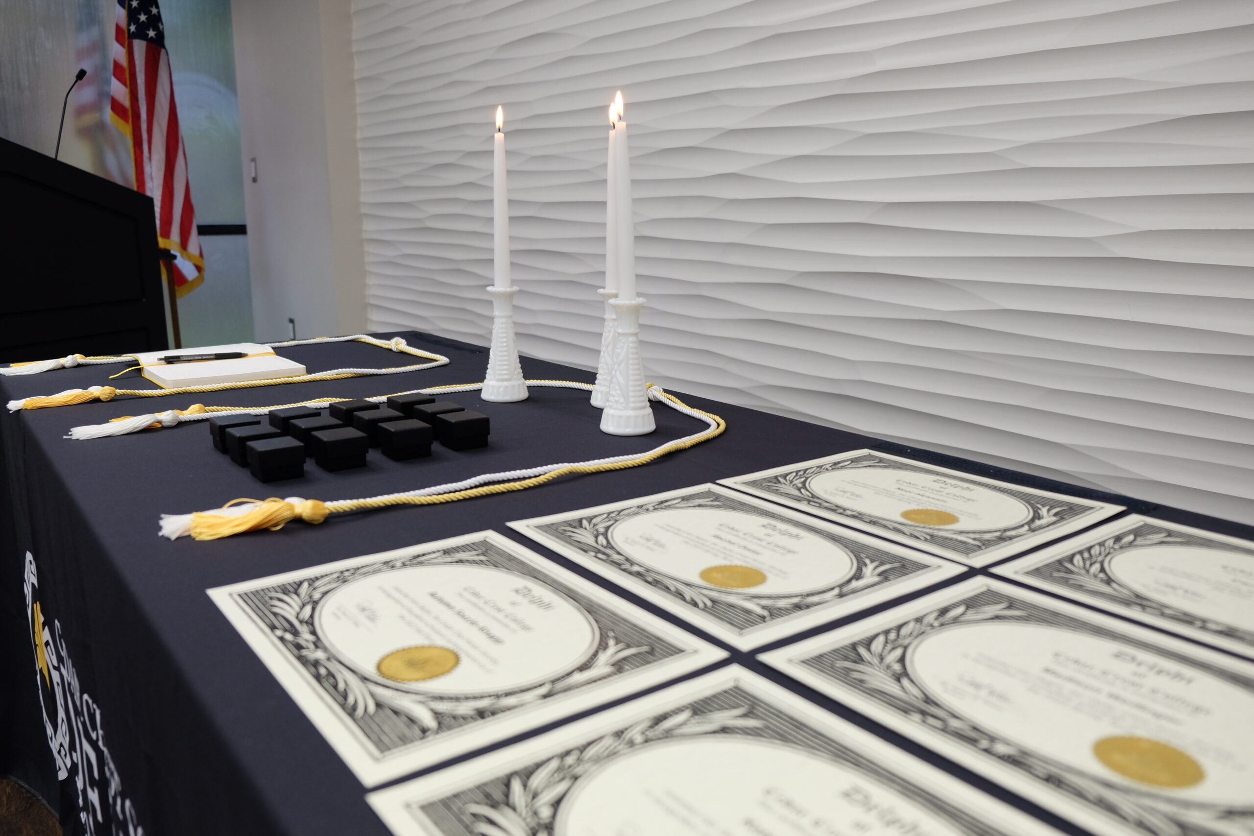 A table with 6 Delphi Honor Society Certificates, three tall, lit candles, a notebook, and two gold and white honors cords laid atop it.