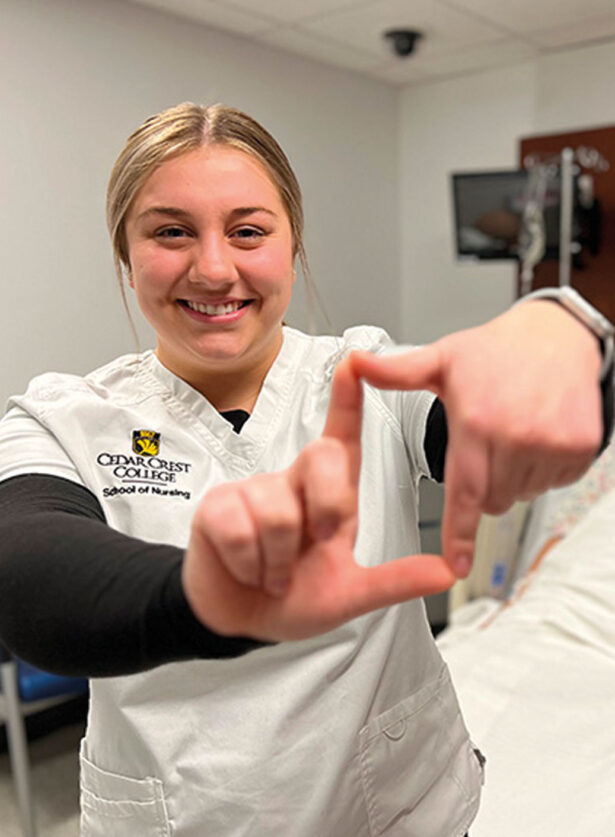 A young, blonde woman wearing a Cedar Crest College School of Nursing uniform, smiling, looking straight ahead. She is holding up her hands, making an 