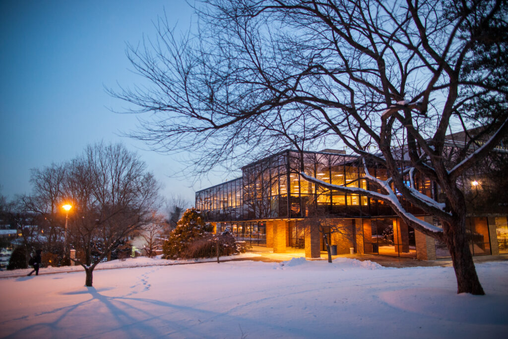 Blaney Hall in the evening, snow covers the campus and lines the bare tree. Warm, orange light emanates from inside TCC