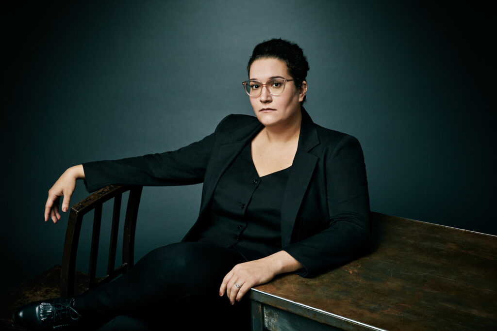 Photo of author Carmen Maria Machado leaning with one arm draped over a chair and another resting on a desk