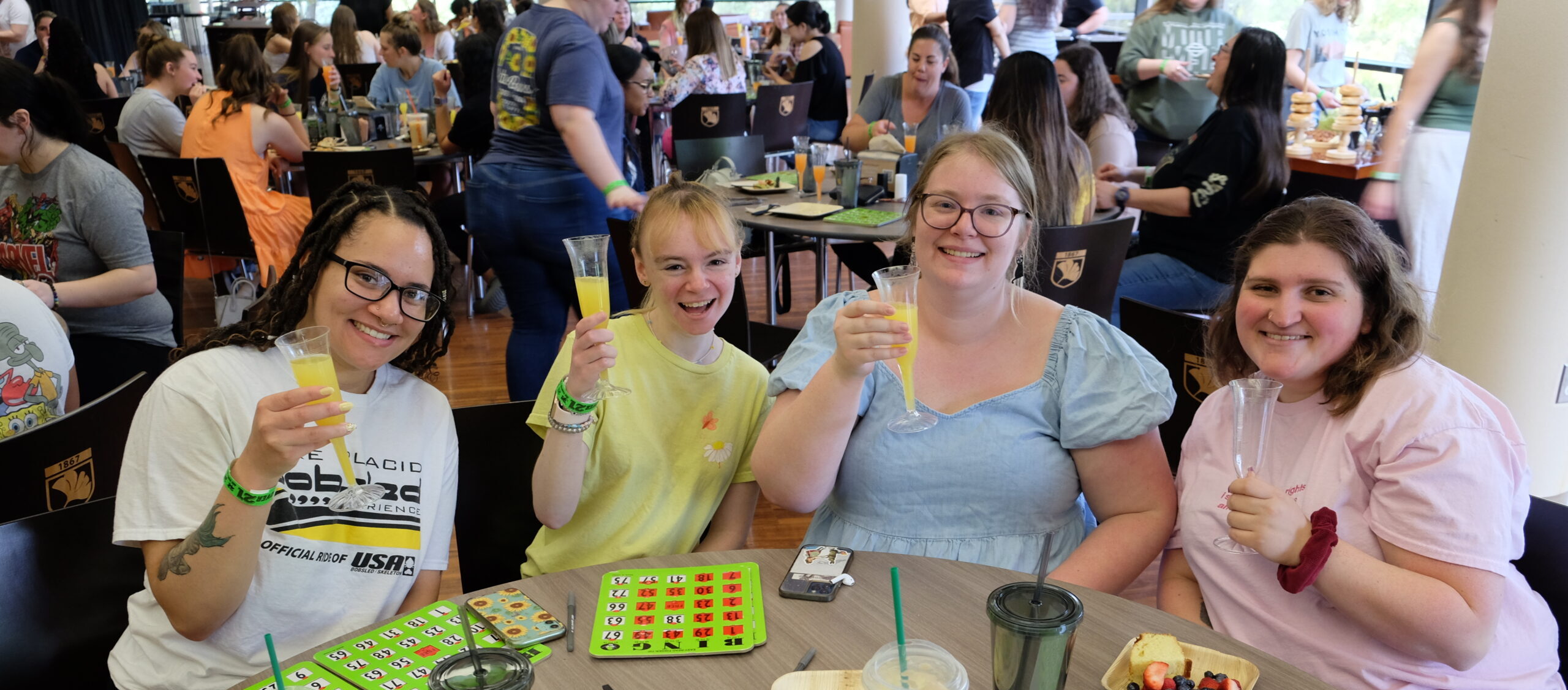 Several students sit around a table with bingo boards and food in front of them, smiles on their faces as they raise their drinks