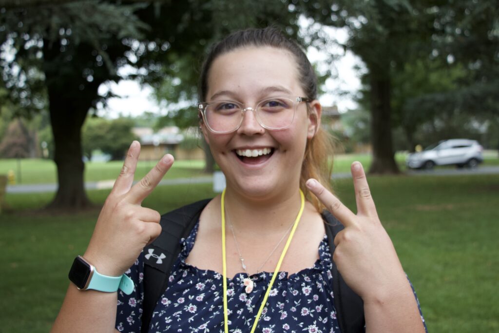 Photo depicts a young girl smiling with her hands in the shape of dual peace signs