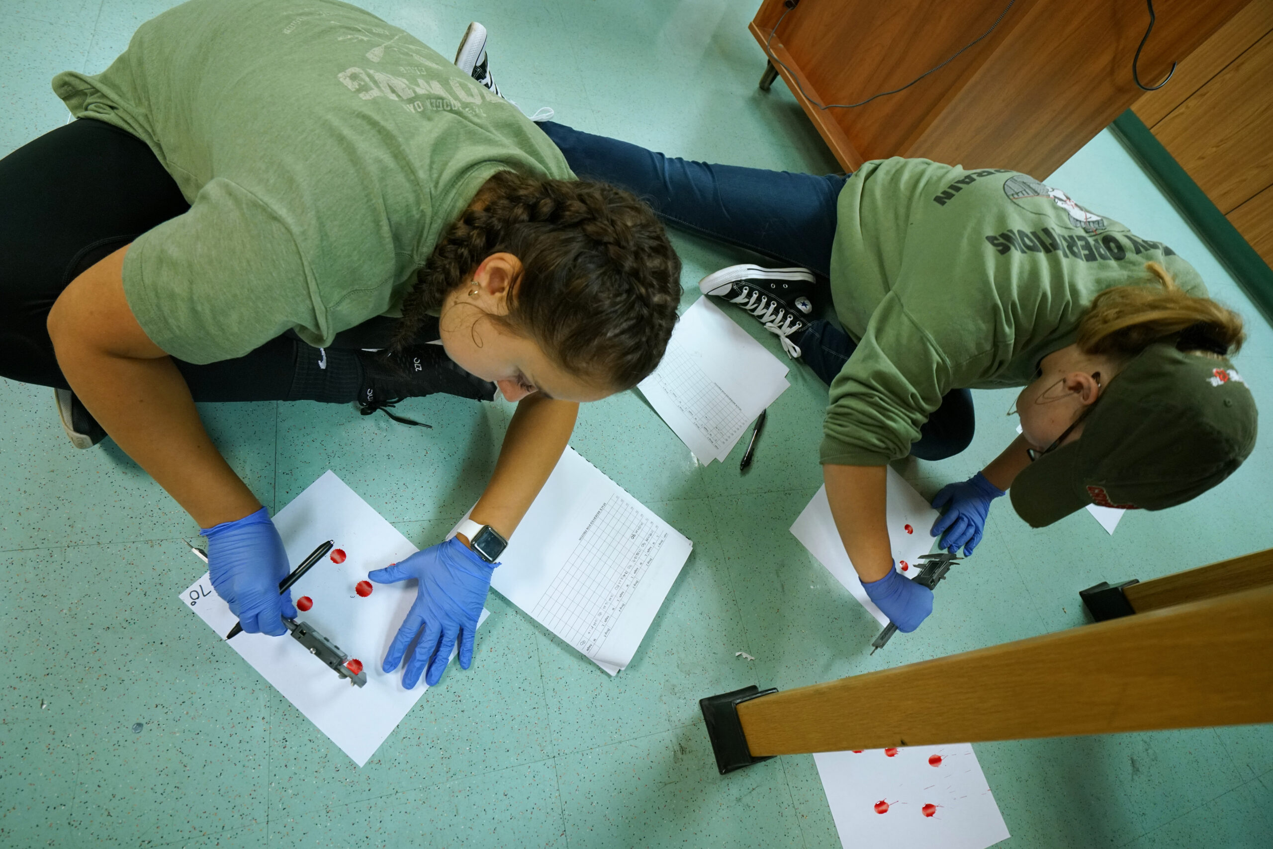 Two girls in green shirts measuring blood splatters on some sheets of paper