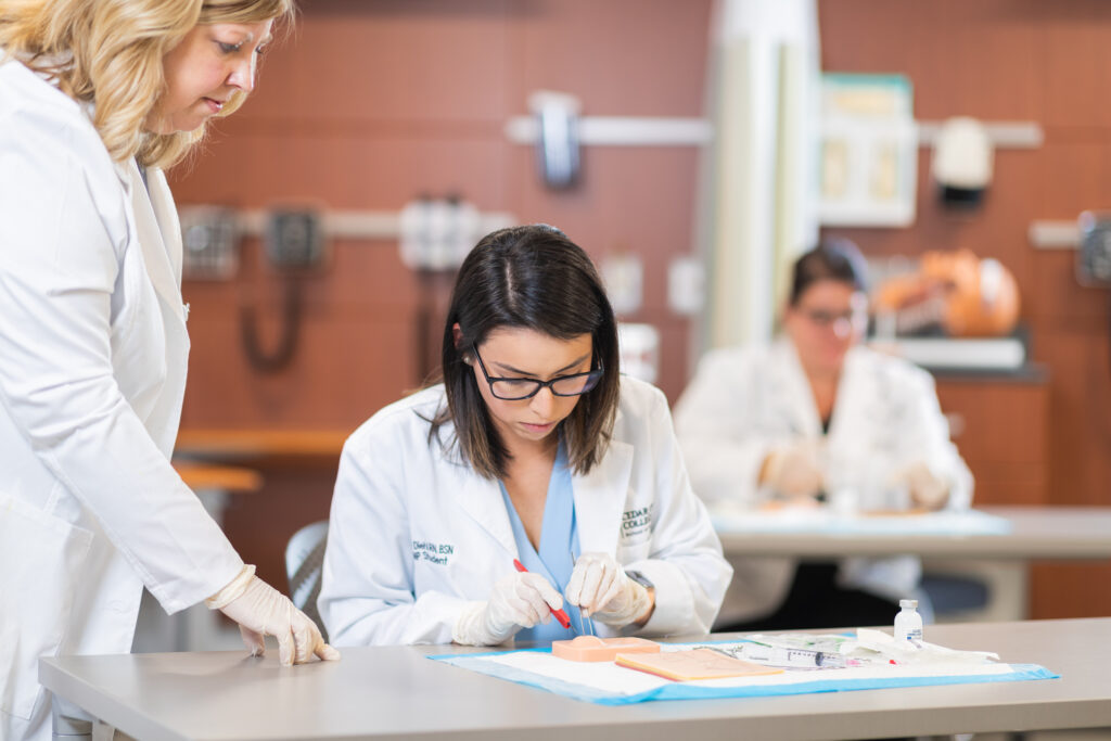 A girl with dark hair and glasses in a white lab coat practicing sutures as a professor in a white lab coat observes