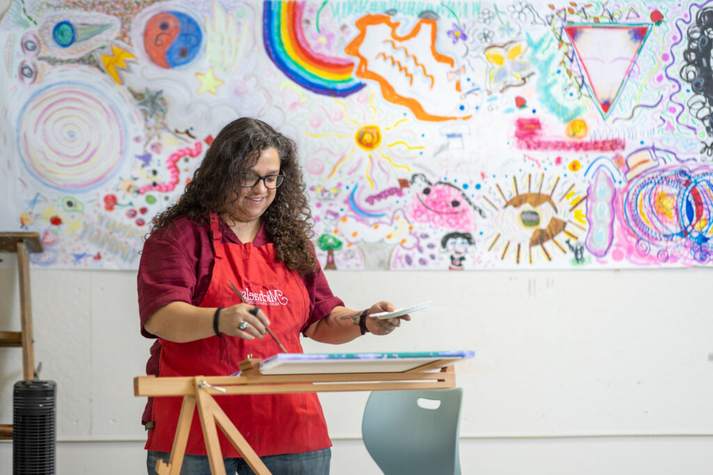 A woman with curly hair and a red shirt and apron smiling as she paints a canvas