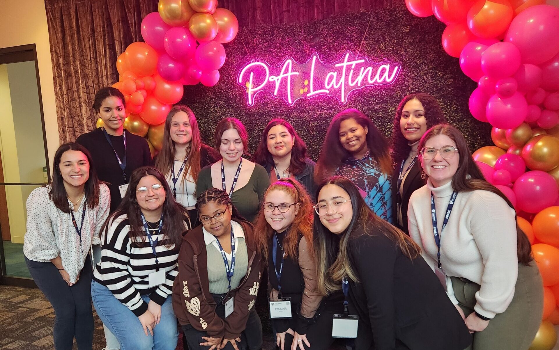 A group of students and staff standing in front of a pink balloon arch and a neon sign that says PA latina